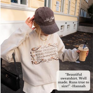 Puede incluir: Una sudadera beige con el texto "WHAT EXCELLENT BOILED POTATOES Such an Exemplary Vegetable Est. 1813" en marr&oacute;n y dorado. La imagen tambi&eacute;n muestra una gorra de b&eacute;isbol marr&oacute;n con "La Paris" en blanco, un bolso negro y una bebida en un vaso transparente.