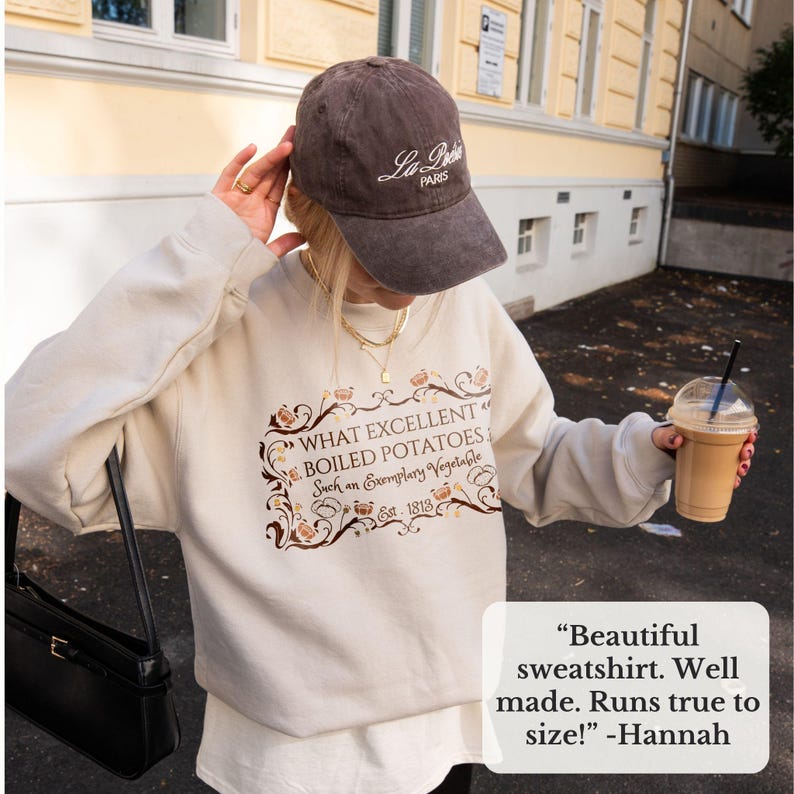 Puede incluir: Una sudadera beige con el texto "WHAT EXCELLENT BOILED POTATOES Such an Exemplary Vegetable Est. 1813" en marr&oacute;n y dorado. La imagen tambi&eacute;n muestra una gorra de b&eacute;isbol marr&oacute;n con "La Paris" en blanco, un bolso negro y una bebida en un vaso transparente.