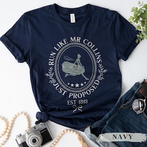 Puede incluir: Camiseta azul marino con un gráfico blanco de una mujer corriendo delante de un hombre con el texto "Run Like Mr. Collins Just Proposed Est 1813".