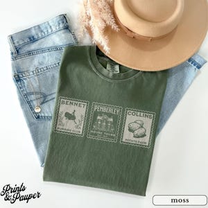 K&ouml;nnte beinhalten: Ein olivgr&uuml;nes T-Shirt mit drei Briefmarken-Designs mit den W&ouml;rtern "Bennet", "Pemberley" und "Collins". Das Shirt wird mit hellblauen Jeans, einer goldenen Halskette und einem beigefarbenen Hut kombiniert. Der Text "Prints & Pauper" steht unten.