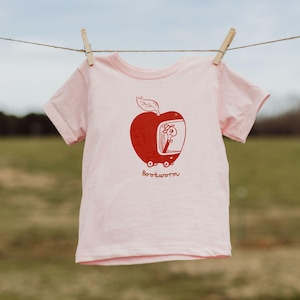 Op de afbeelding: Lichtroze T-shirt met een rode afbeelding van een appel met een worm erin en het woord "Bookworm" eronder. Het T-shirt hangt aan een waslijn met houten knijpers.
