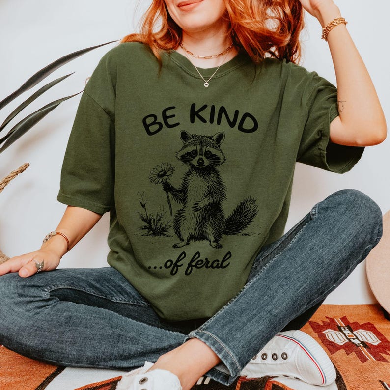 May include: A green t-shirt with a black graphic of a raccoon holding a flower and the text "Be Kind... of feral".