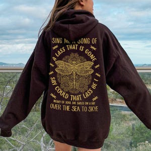 May include: Dark brown hoodie featuring a gold dragonfly design and text. The text reads "Sing me a song of a lass that is gone, say could that lass be I, merry of soul she sailed on a day over the sea to Skye."