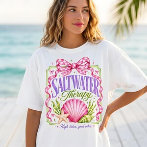 Könnte beinhalten: Weißes T-Shirt mit einem farbenfrohen Grafikdesign. Das Design zeigt die Worte "SALTWATER Therapy" in lila Schrift, eine rosa Muschel, einen Seestern und eine karierte rosa Schleife. Der Text "High tides, good vibes" ist ebenfalls enthalten.