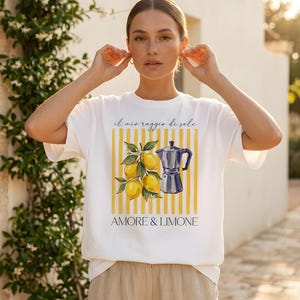 May include: A white short-sleeved t-shirt featuring a graphic print of yellow lemons and a blue moka pot over yellow vertical stripes. The text "il mio raggio di sole" and "AMORE & LIMONE" is printed in black above and below the graphic design.