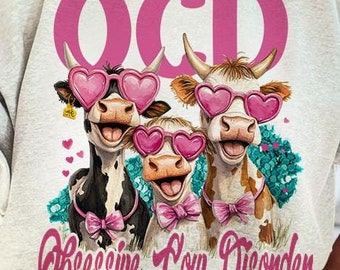 May include: A white sweatshirt with a pink graphic of three cows wearing heart-shaped sunglasses. The text "OCD Obsessive Cow Disorder" is printed below the cows.