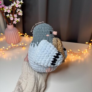 Pu&ograve; includere: Un giocattolo di peluche a forma di piccione all'uncinetto con la testa grigia e il corpo blu. Il piccione tiene un oggetto marrone all'uncinetto nella sua ala. Il giocattolo &egrave; tenuto da una mano che indossa un maglione beige.