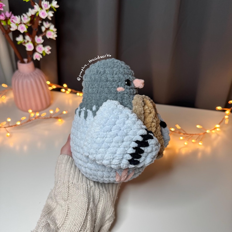 Pu&ograve; includere: Un giocattolo di peluche a forma di piccione all'uncinetto con la testa grigia e il corpo blu. Il piccione tiene un oggetto marrone all'uncinetto nella sua ala. Il giocattolo &egrave; tenuto da una mano che indossa un maglione beige.