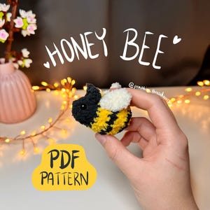 Puede incluir: Una abeja de peluche hecha a mano con detalles en negro, amarillo y blanco. El texto "HONEY BEE" está escrito encima. También es visible un letrero amarillo con "PDF PATTERN". La abeja está sostenida en una mano.