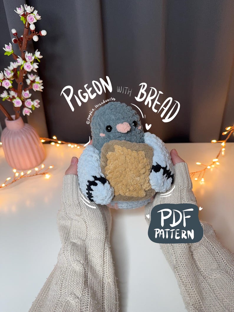 Pu&ograve; includere: Un piccione amigurumi all'uncinetto che tiene un pezzo di pane. Il piccione &egrave; grigio con un becco rosa e occhi neri. Il pane &egrave; marrone e ha una crosta bianca. Il testo "PIGEON WITH BREAD" &egrave; scritto in bianco sopra il piccione. Il testo "PDF PATTERN" &egrave; scritto in bianco sotto il piccione.