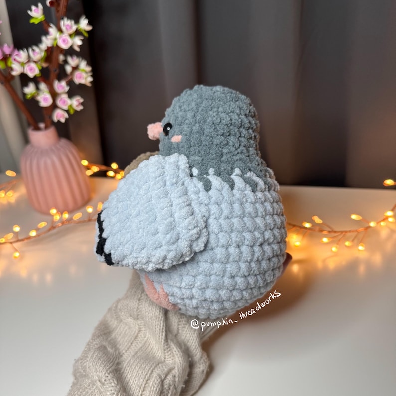 Pu&ograve; includere: Un giocattolo di peluche all'uncinetto a forma di colomba. La colomba &egrave; di colore blu chiaro e grigio con un becco rosa e si trova su un tessuto di maglia beige. La colomba &egrave; rivolta verso la parte posteriore della fotocamera.