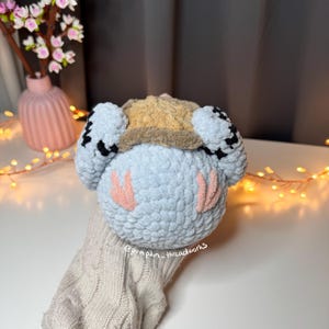 Pu&ograve; includere: Un giocattolo di peluche all'uncinetto con un corpo blu, guance rosa e un cappello a scacchi marrone e nero. Il giocattolo &egrave; seduto su una superficie a maglia.