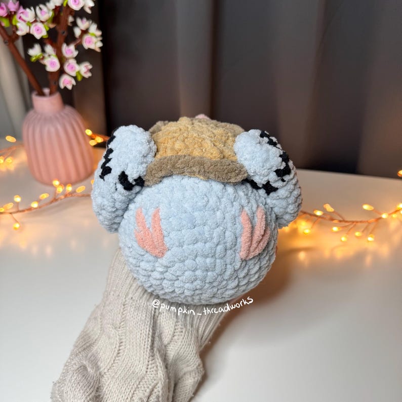 Pu&ograve; includere: Un giocattolo di peluche all'uncinetto con un corpo blu, guance rosa e un cappello a scacchi marrone e nero. Il giocattolo &egrave; seduto su una superficie a maglia.
