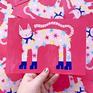 May include: Pink cat art print with a blue scarf and star cutouts in red, yellow, and blue. The cat has blue boots and a striped tail. The background is a solid pink color.