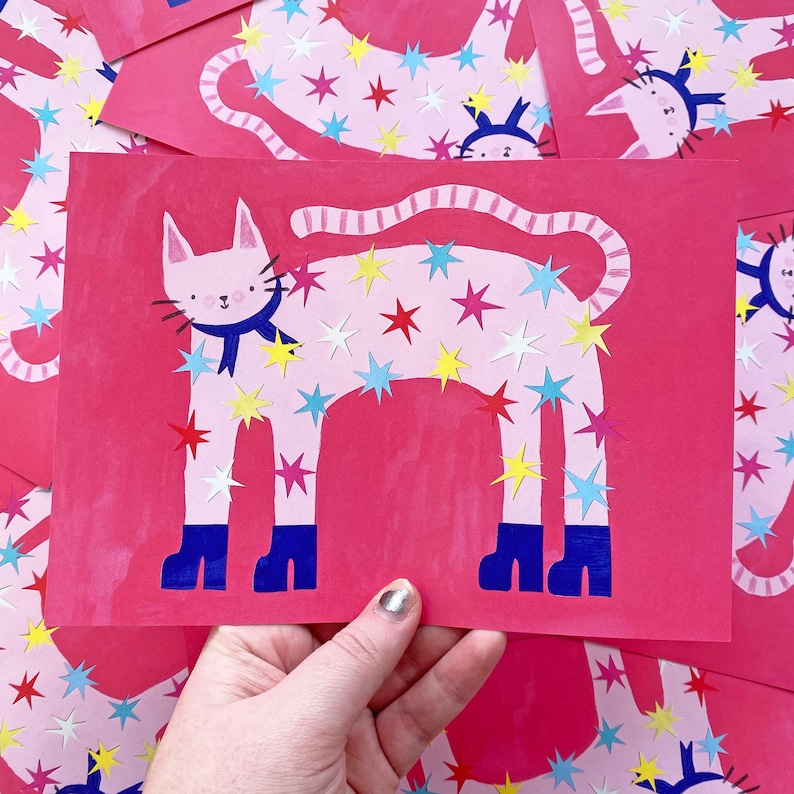 May include: Pink cat art print with a blue scarf and star cutouts in red, yellow, and blue. The cat has blue boots and a striped tail. The background is a solid pink color.