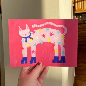 May include: A pink cat illustration with a blue scarf and blue boots, adorned with colorful star cutouts. The cat is set against a bright pink background. The artwork is on a rectangular card.