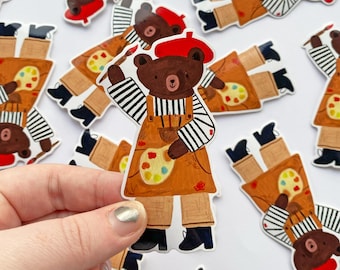 May include: A collection of stickers featuring a brown bear artist wearing a red beret, striped shirt, and brown overalls. The bear holds a paintbrush and a palette with colorful paint. The stickers are on a white background.