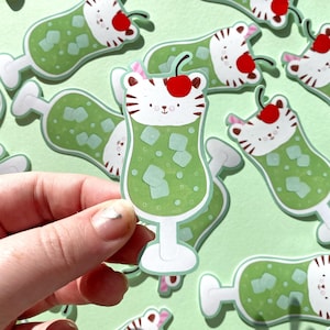May include: A sticker featuring a cartoon cocktail with a tiger face, green drink, and cherry on top. The sticker has a white and green color scheme with a cute design. The sticker is held by a hand.