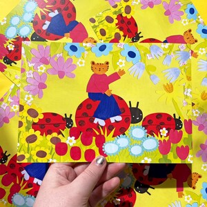 May include: A whimsical illustration on yellow paper features a tiger character riding a ladybug. The scene is surrounded by colorful flowers and more ladybugs. The artwork has a playful, childlike aesthetic.