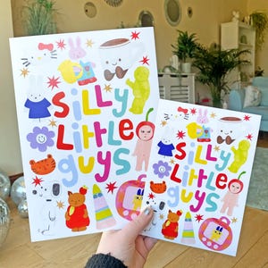 May include: Two colorful prints with the text "Silly Little Guys" and illustrations of cartoon characters. The prints are on white paper with a white background.