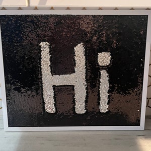 Może przedstawiać: Poduszka w ramie z czarnych i srebrnych cekinów z napisem "Hi" w kolorze białym.