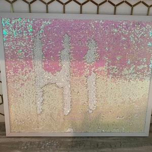 May include: A white framed picture with a pink and white iridescent sequin background. The sequins are arranged to form the word "Hi" in white.