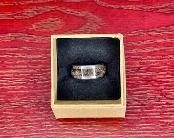 May include: A silver-toned ring with a textured, dark brown center band is displayed in a small, open cardboard box with a black interior. The ring has a polished, metallic finish and a unique, speckled design.
