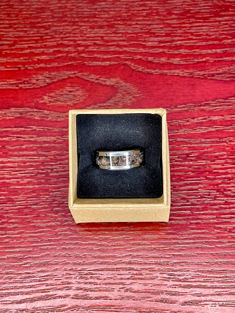 K&ouml;nnte beinhalten: Ein silberfarbener Ring mit einem strukturierten, dunkelbraunen Mittelband wird in einer kleinen, offenen Pappschachtel mit schwarzem Innenraum pr&auml;sentiert. Der Ring hat eine polierte Metalloberfl&auml;che und ein einzigartiges, gesprenkeltes Design.