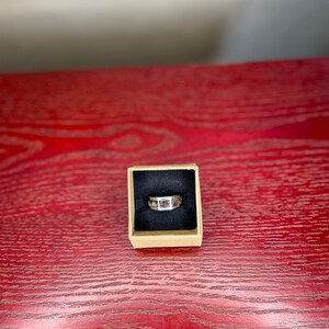 K&ouml;nnte beinhalten: Ein silberner Ring mit einem Band, das ein Mosaik aus braunen und beigefarbenen Steinen aufweist, pr&auml;sentiert in einer kleinen, quadratischen Pappschachtel mit schwarzem Innenraum. Der Ring liegt auf einer roten Holzoberfl&auml;che.