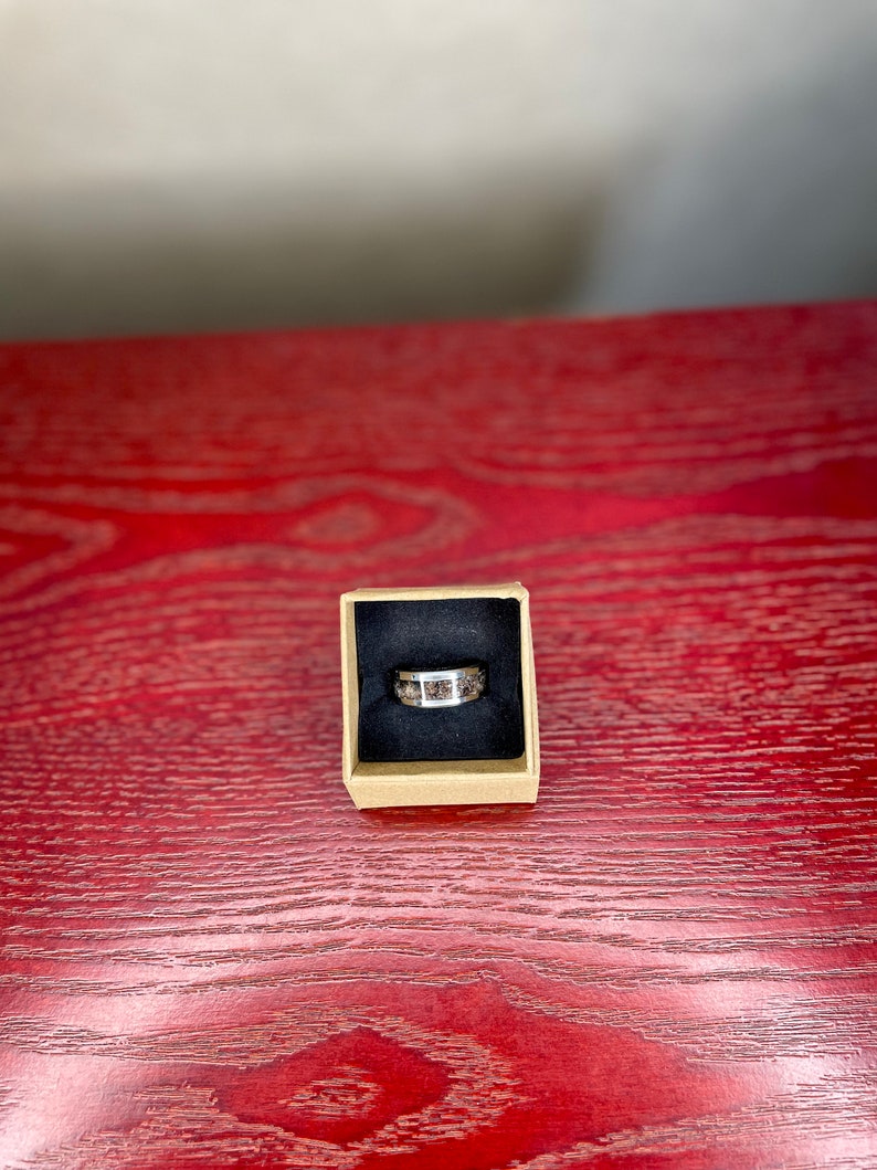 K&ouml;nnte beinhalten: Ein silberner Ring mit einem Band, das ein Mosaik aus braunen und beigefarbenen Steinen aufweist, pr&auml;sentiert in einer kleinen, quadratischen Pappschachtel mit schwarzem Innenraum. Der Ring liegt auf einer roten Holzoberfl&auml;che.