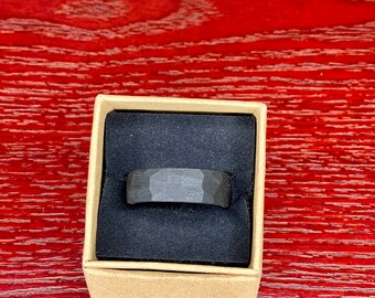 Könnte beinhalten: Ein dunkelgrauer, gehammerter Metallring, der in einer kleinen, offenen Pappschachtel mit schwarzem Innenraum präsentiert wird. Der Ring hat eine strukturierte Oberfläche und ein Banddesign. Die Schachtel steht auf einer roten Holzoberfläche.