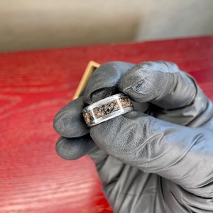 K&ouml;nnte beinhalten: Ein silberner Ring mit einem Band, das einen strukturierten, erdfarbenen Einsatz aufweist. Der Ring wird von einer behandschuhten Hand gehalten, die die kontrastierenden Farben und Texturen des Schmucks zeigt.