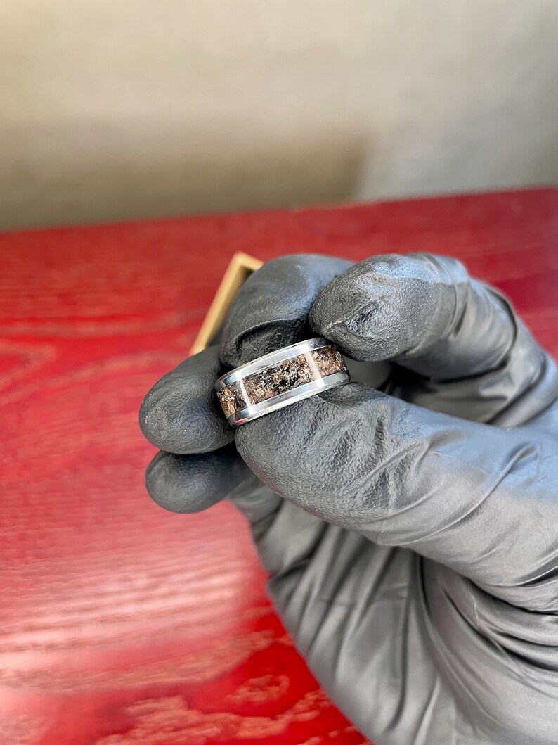 K&ouml;nnte beinhalten: Ein silberner Ring mit einem Band, das einen strukturierten, erdfarbenen Einsatz aufweist. Der Ring wird von einer behandschuhten Hand gehalten, die die kontrastierenden Farben und Texturen des Schmucks zeigt.
