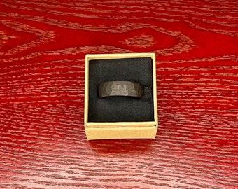 Könnte beinhalten: Ein dunkelgrauer, gehammerter Metallring, der in einer kleinen, offenen Pappschachtel mit schwarzem Innenraum präsentiert wird. Der Ring hat eine strukturierte Oberfläche und eine Bandbreite von ca. 0,6 cm. Die Schachtel steht auf einer rot getönten Holzoberfläche.