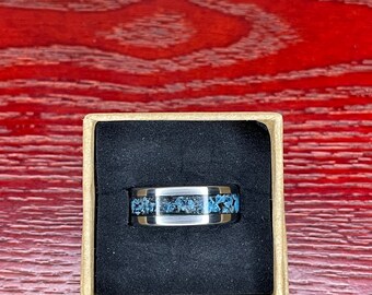 Könnte beinhalten: Ein silberfarbener Ring mit dunkler Einlage, die blaue und graue Steinsplitter enthält. Der Ring wird in einer kleinen, offenen Pappschachtel mit schwarzem Innenraum auf einer rotbraunen Holzoberfläche präsentiert.