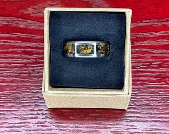 Könnte beinhalten: Ein Edelstahlring mit einem rechteckigen Einsatz aus gold-braunem Stein. Der Ring wird in einer kleinen, offenen Pappschachtel mit schwarzem Innenraum auf einer roten Holzmaserung präsentiert.