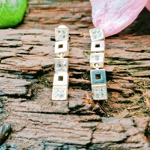 Puede incluir: Un par de pendientes colgantes plateados. Cada pendiente presenta una serie de formas cuadradas y rectangulares, algunas adornadas con pequeños cristales transparentes. Los pendientes se exhiben sobre una superficie de madera marrón texturizada.