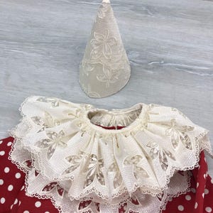 May include: A cream-colored, lace-trimmed clown collar and matching hat. The collar is layered with a ruffled edge and floral cutouts. The hat is cone-shaped with a floral pattern. The outfit is set against a red polka-dot background.