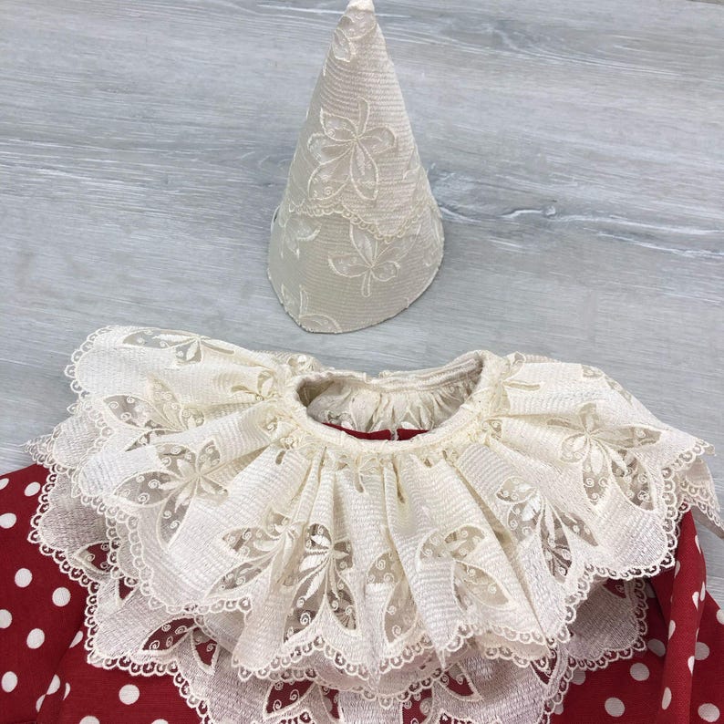 May include: A cream-colored, lace-trimmed clown collar and matching hat. The collar is layered with a ruffled edge and floral cutouts. The hat is cone-shaped with a floral pattern. The outfit is set against a red polka-dot background.