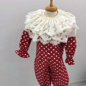 May include: A red and white polka dot clown costume with a large, layered white lace collar. The long-sleeved jumpsuit has ruffled cuffs and ankles. The costume is displayed on a mannequin.