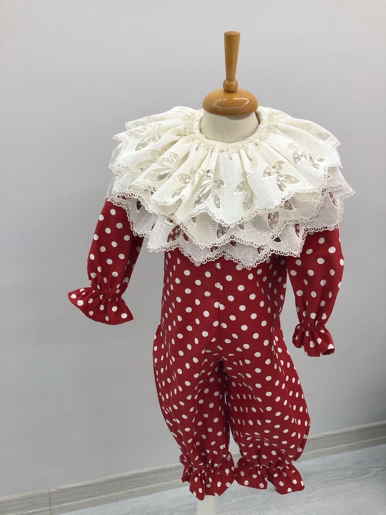May include: A red and white polka dot clown costume with a large, layered white lace collar. The long-sleeved jumpsuit has ruffled cuffs and ankles. The costume is displayed on a mannequin.