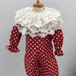 May include: A red and white polka dot clown costume with a large, layered lace collar. The long-sleeved jumpsuit has ruffled cuffs and ankles. The costume is displayed on a mannequin.