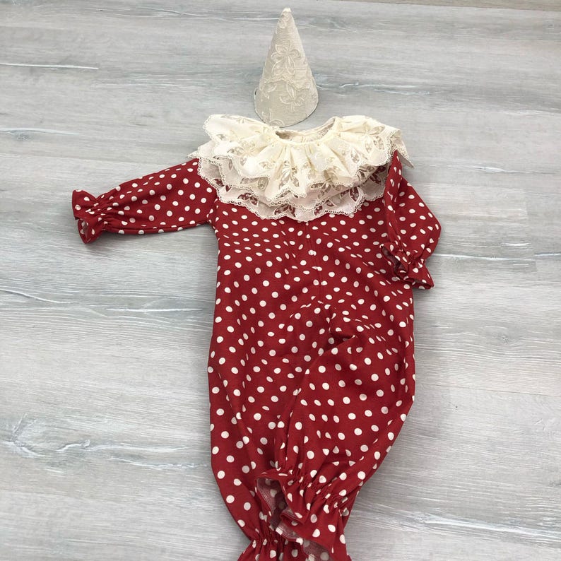 May include: A red and white polka dot baby outfit with a cream-colored lace collar and matching hat. The long-sleeved outfit has a ruffled hem and cuffs. The hat is cone-shaped and made of a cream-colored lace fabric.