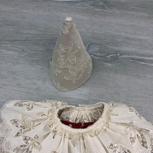 May include: A cream-colored, vintage-style lace collar and matching conical hat. The collar features a ruffled design with floral cutouts and a red interior. The hat is beige with a floral lace pattern. The items appear to be part of a historical costume.
