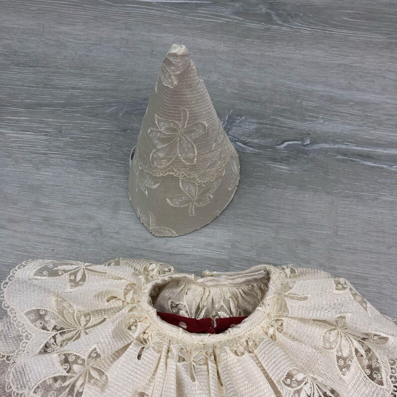 May include: A cream-colored, vintage-style lace collar and matching conical hat. The collar features a ruffled design with floral cutouts and a red interior. The hat is beige with a floral lace pattern. The items appear to be part of a historical costume.
