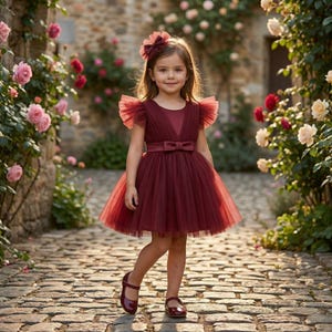 May include: A young girl in a burgundy tulle dress with a matching bow and shoes stands on a cobblestone path. The dress features a V-neck, flutter sleeves, and a bow at the waist. The background includes a stone wall and blooming roses.