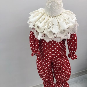 May include: A red and white polka dot clown costume with a ruffled collar and matching hat. The long-sleeved jumpsuit has white polka dots and ruffled cuffs at the wrists and ankles. The hat and collar are white with a floral lace pattern.