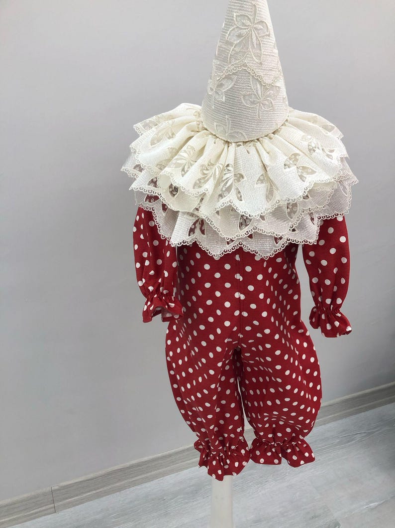 May include: A red and white polka dot clown costume with a ruffled collar and matching hat. The long-sleeved jumpsuit has white polka dots and ruffled cuffs at the wrists and ankles. The hat and collar are white with a floral lace pattern.