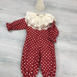May include: A red and white polka dot clown costume with a ruffled collar and matching hat. The long-sleeved jumpsuit has white polka dots and ruffled cuffs at the ankles and wrists. The hat is cone-shaped and made of a cream-colored lace fabric.