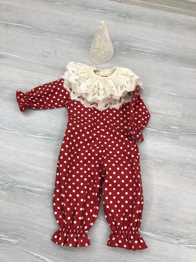May include: A red and white polka dot clown costume with a ruffled collar and matching hat. The long-sleeved jumpsuit has white polka dots and ruffled cuffs at the ankles and wrists. The hat is cone-shaped and made of a cream-colored lace fabric.
